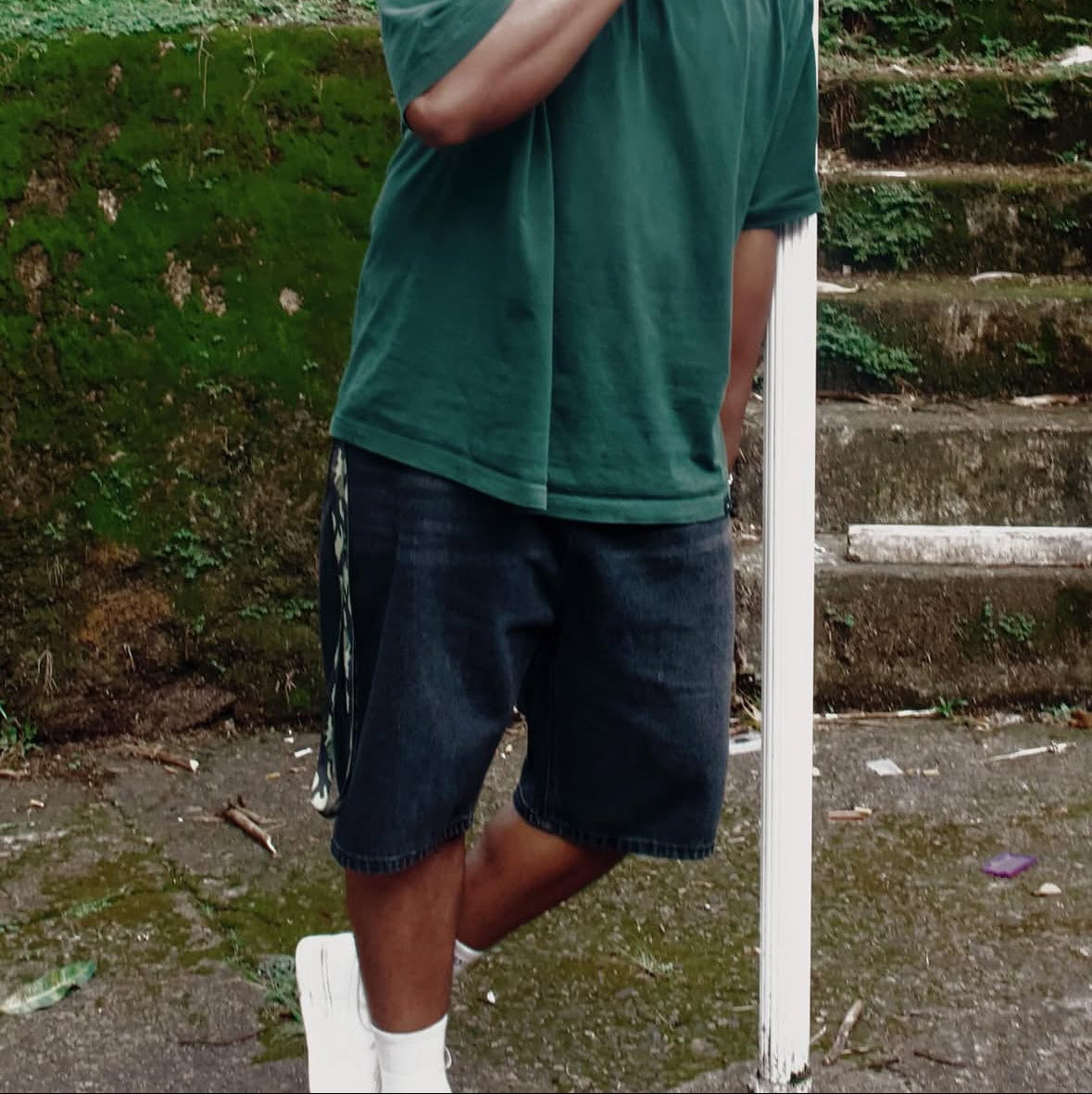 Person wearing a green shirt and dark shorts standing on a concrete surface with a white pole and green wall in the background.