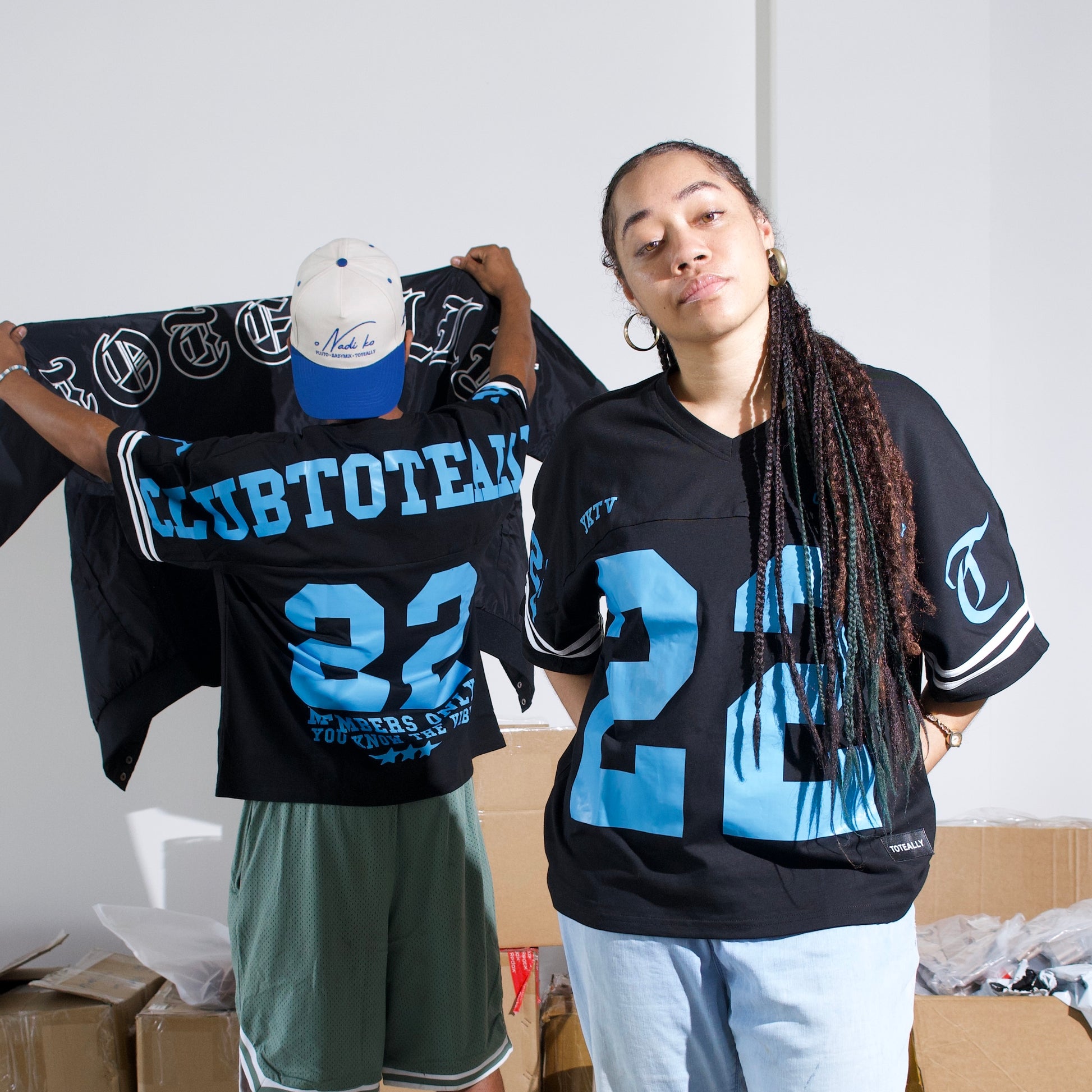 Two people wearing sports jerseys with numbers and text, standing in a room with boxes and bags.