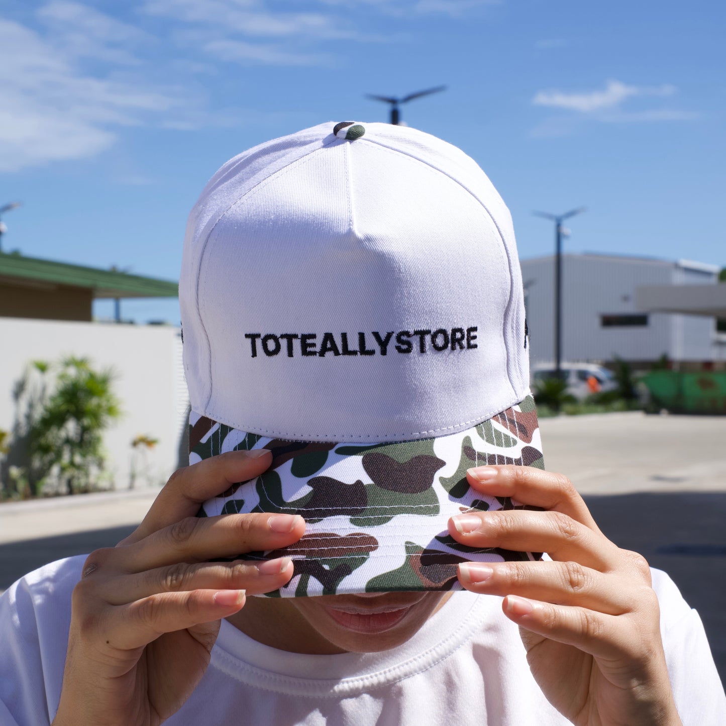 Person holding a camouflage phone case with a white cap labeled 'TOTALLY STORE' against a blue sky.
