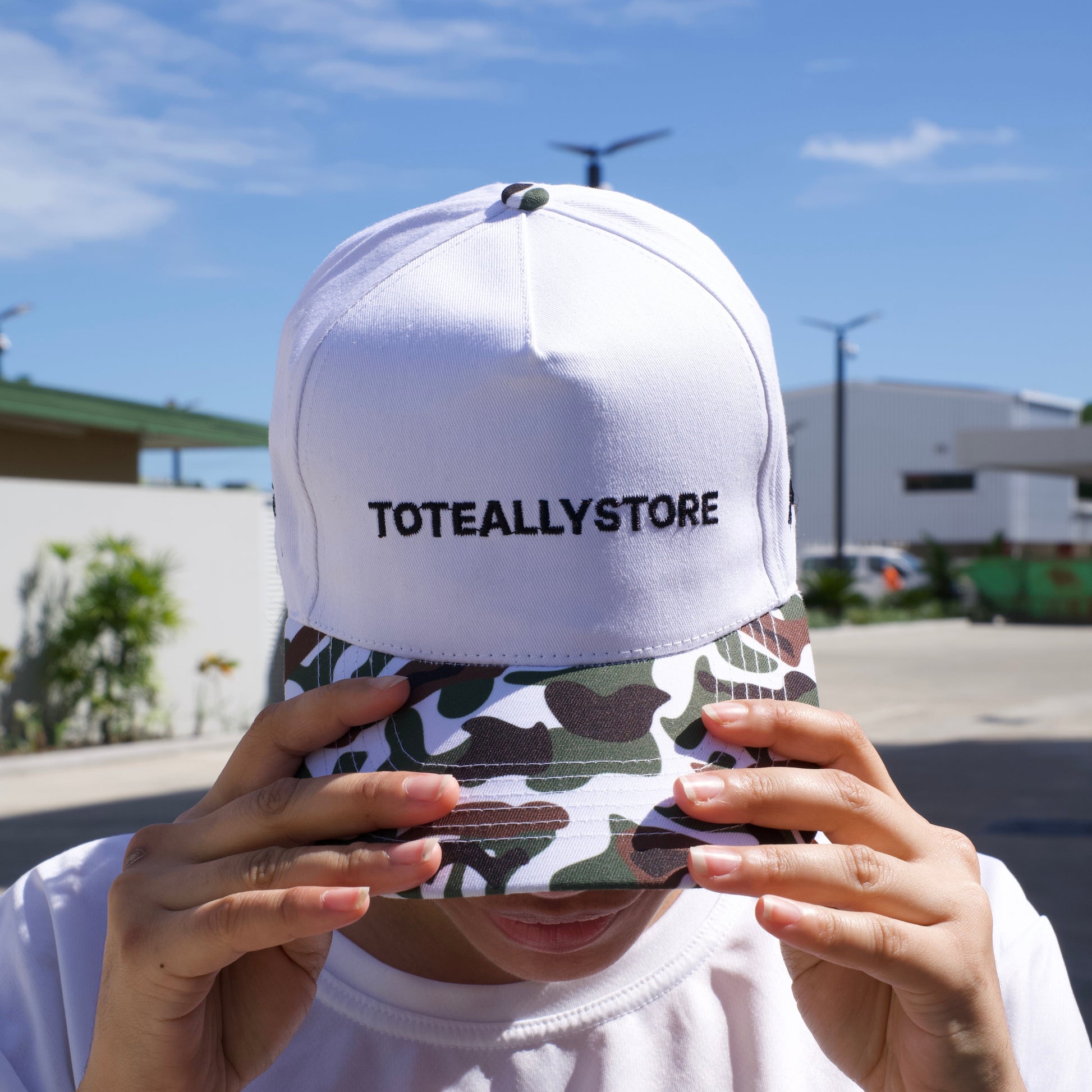 Person holding a camouflage phone case with a white cap labeled 'TOTALLY STORE' against a blue sky.