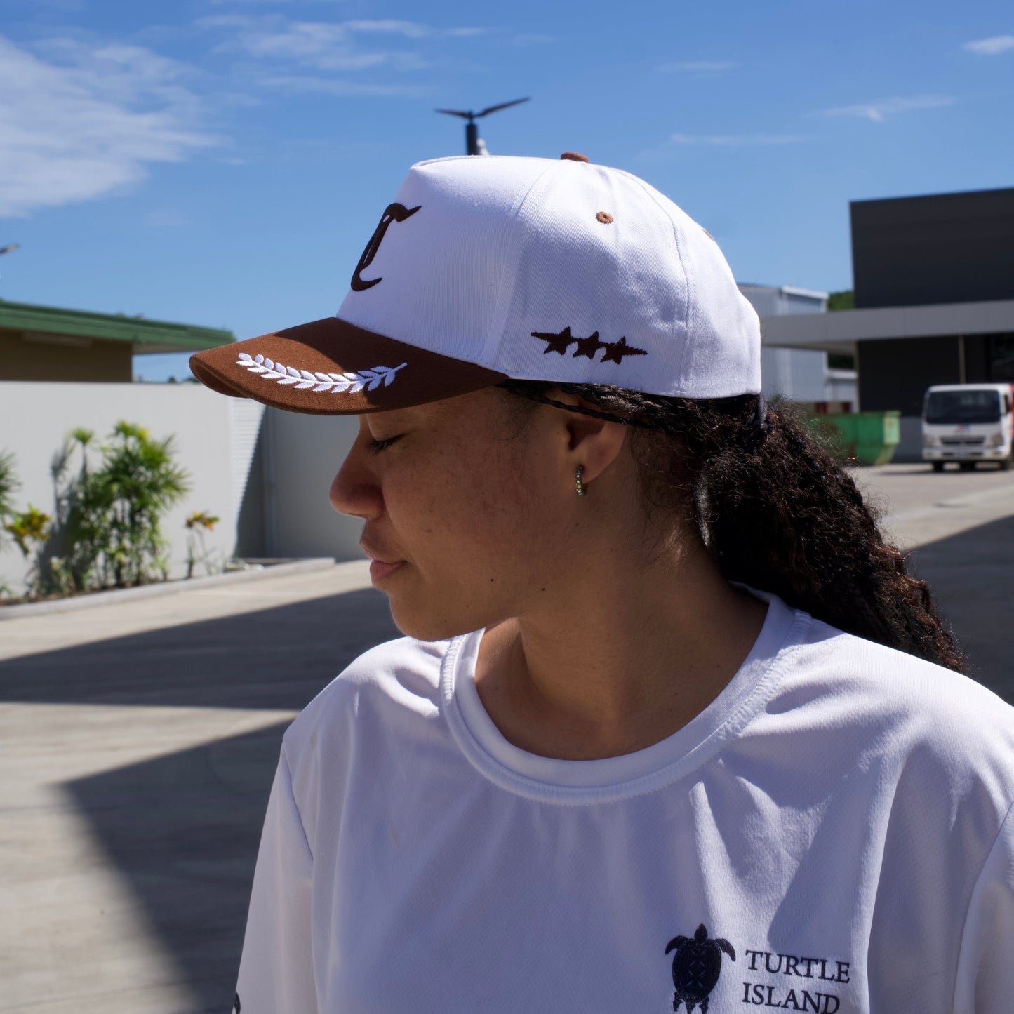 Person wearing a white tank top with a logo and a cap, standing outdoors with a clear blue sky.
