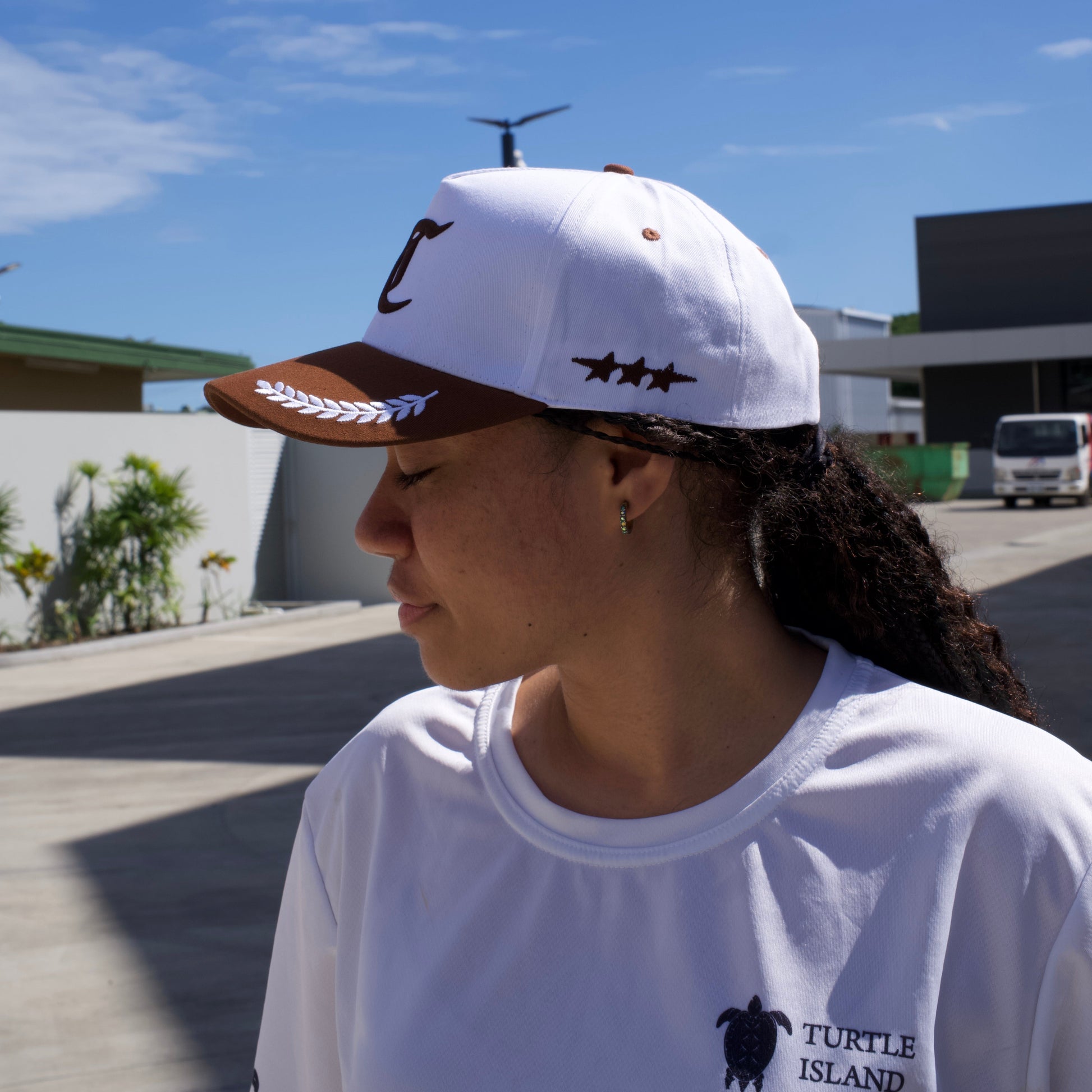 Person wearing a white tank top with a logo and a cap, standing outdoors with a clear blue sky.