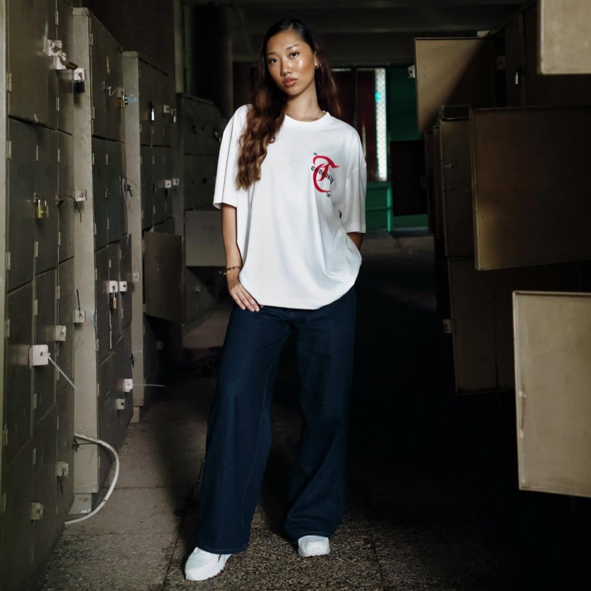 Person wearing a white t-shirt with a red logo in a dimly lit room.