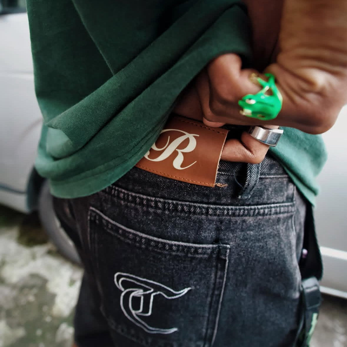 Person wearing a green hoodie and dark jeans with a brand logo, standing next to a white car.