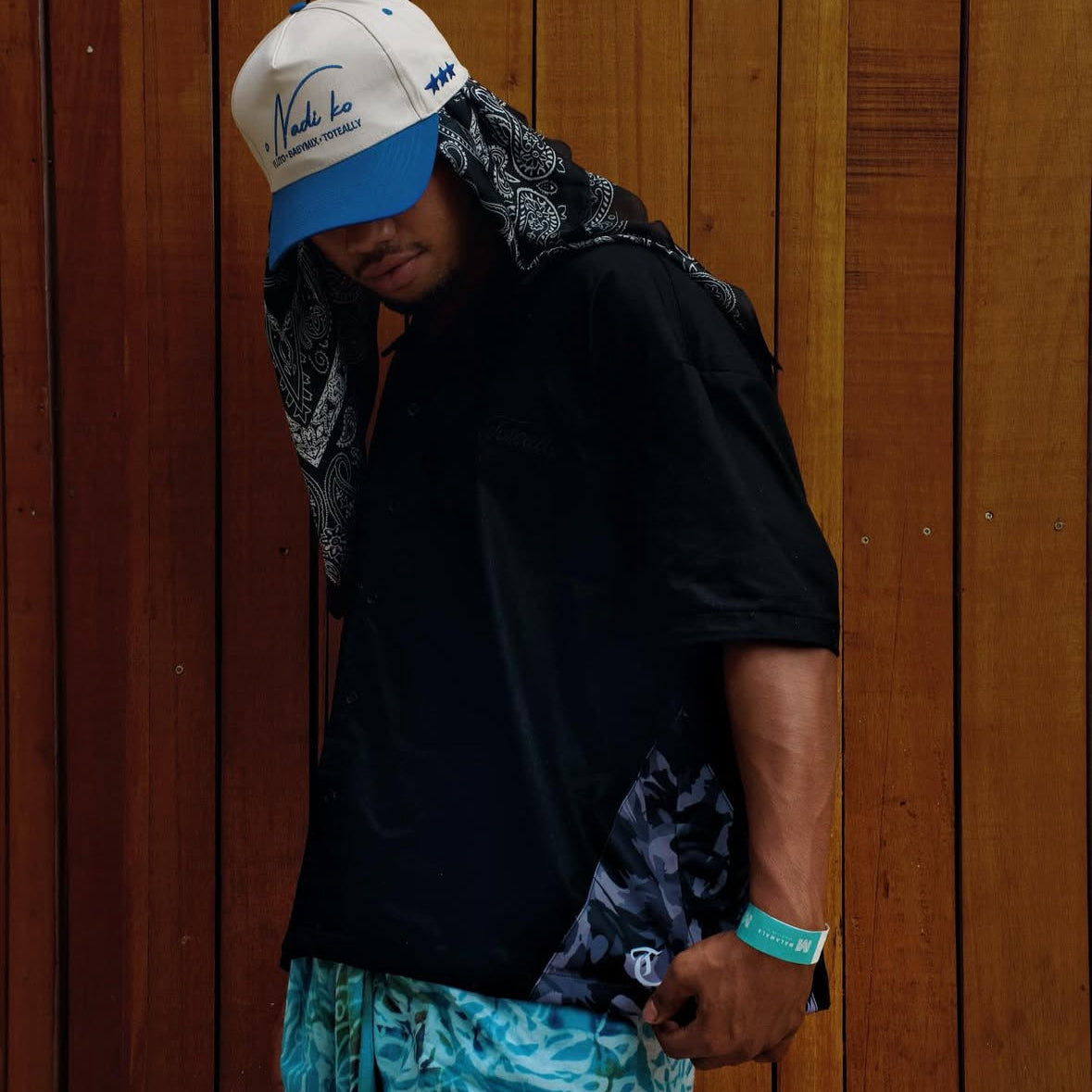 Person wearing a black shirt, blue shorts, and a white cap with a blue bill against a wooden wall.