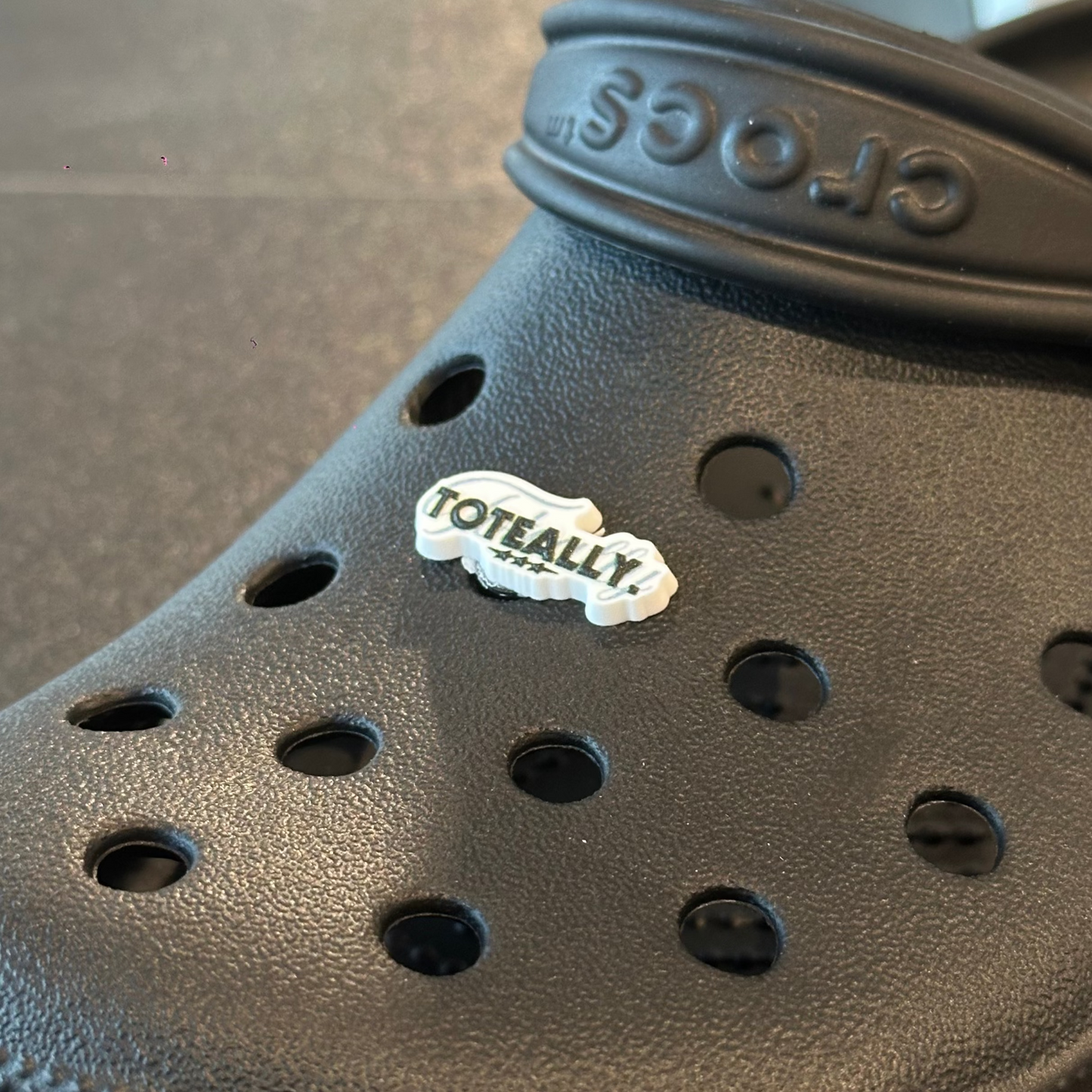 Black croc shoe with a 'TOTALLY!' sticker on a gray surface